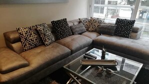 A sectional sofa with patterned cushions, a glass coffee table with a metal frame, and a view of the outdoors through a large window.