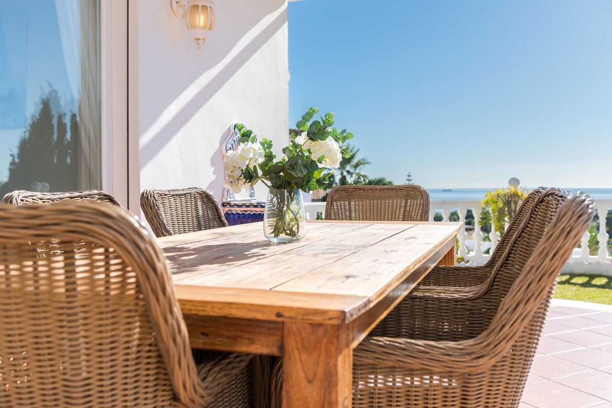 Una mesa de madera con sillas de mimbre, un florero con flores y vistas al mar.