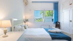 A bedroom with a bed, bedside table, lamp, window with blue curtains, and a radiator. - Villa Panorama – Ocean View Oasis with Private Pool, Mijas Costa (Mijas Costa)