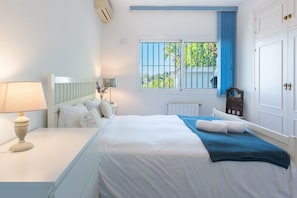 A bedroom with a bed, bedside table, lamp, window with blue curtains, and a radiator. - Villa Panorama – Ocean View Oasis with Private Pool, Mijas Costa (Mijas Costa)