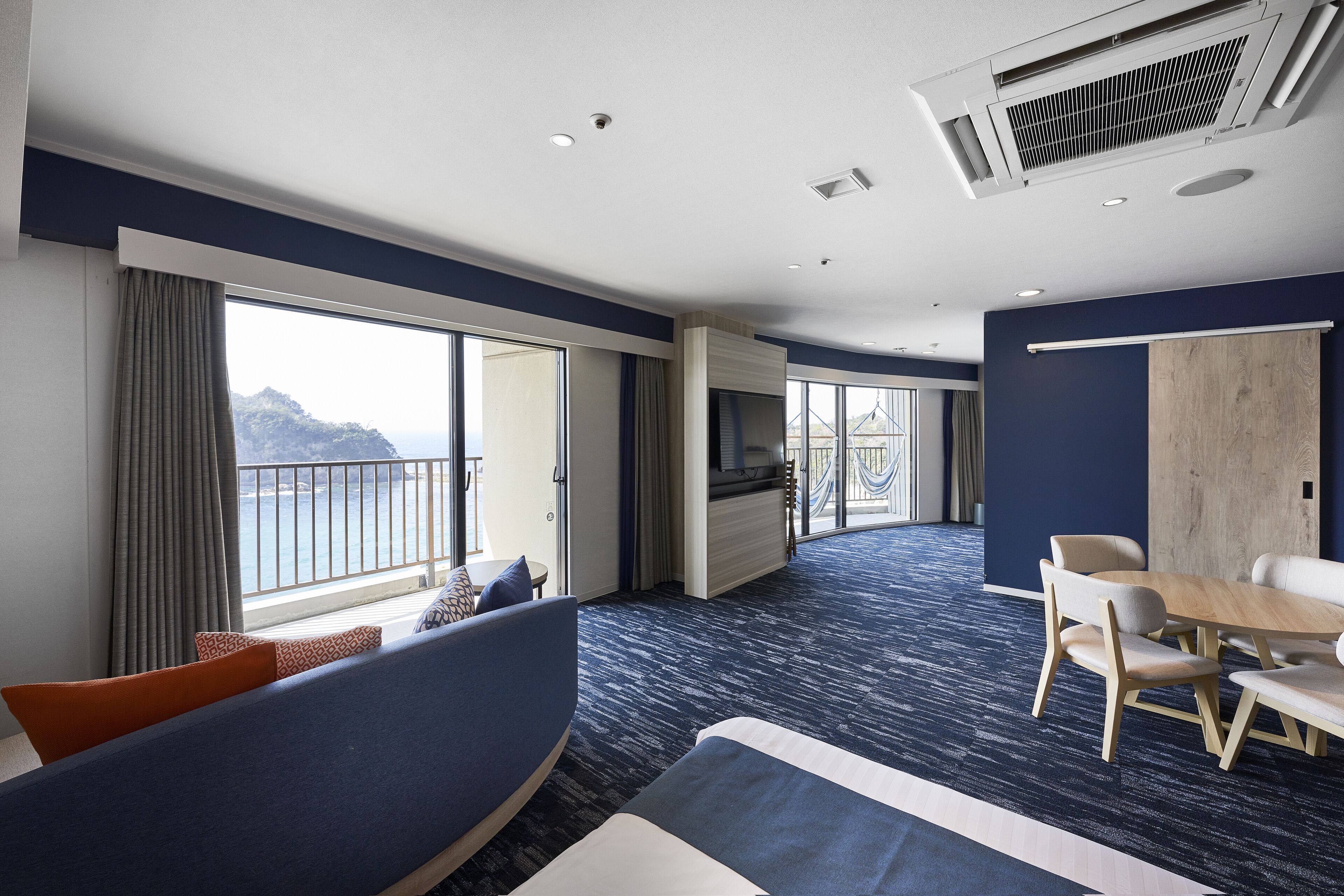 A modern hotel room with a large bed, a dining area, and a balcony with a view of the sea.