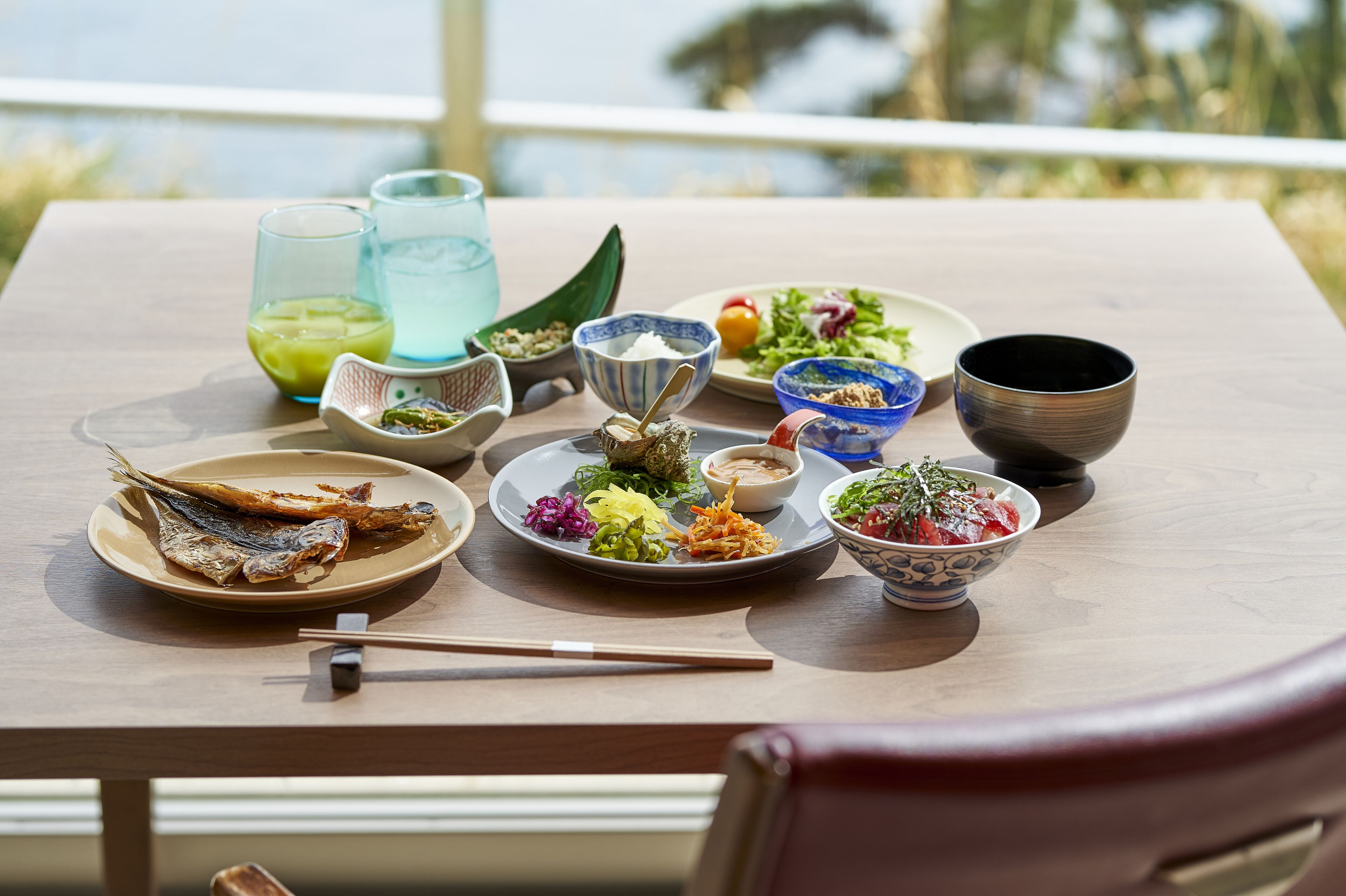 A dining table with various dishes, including fish, salads, and bowls of food, set on a wooden table with a view of the outdoors.