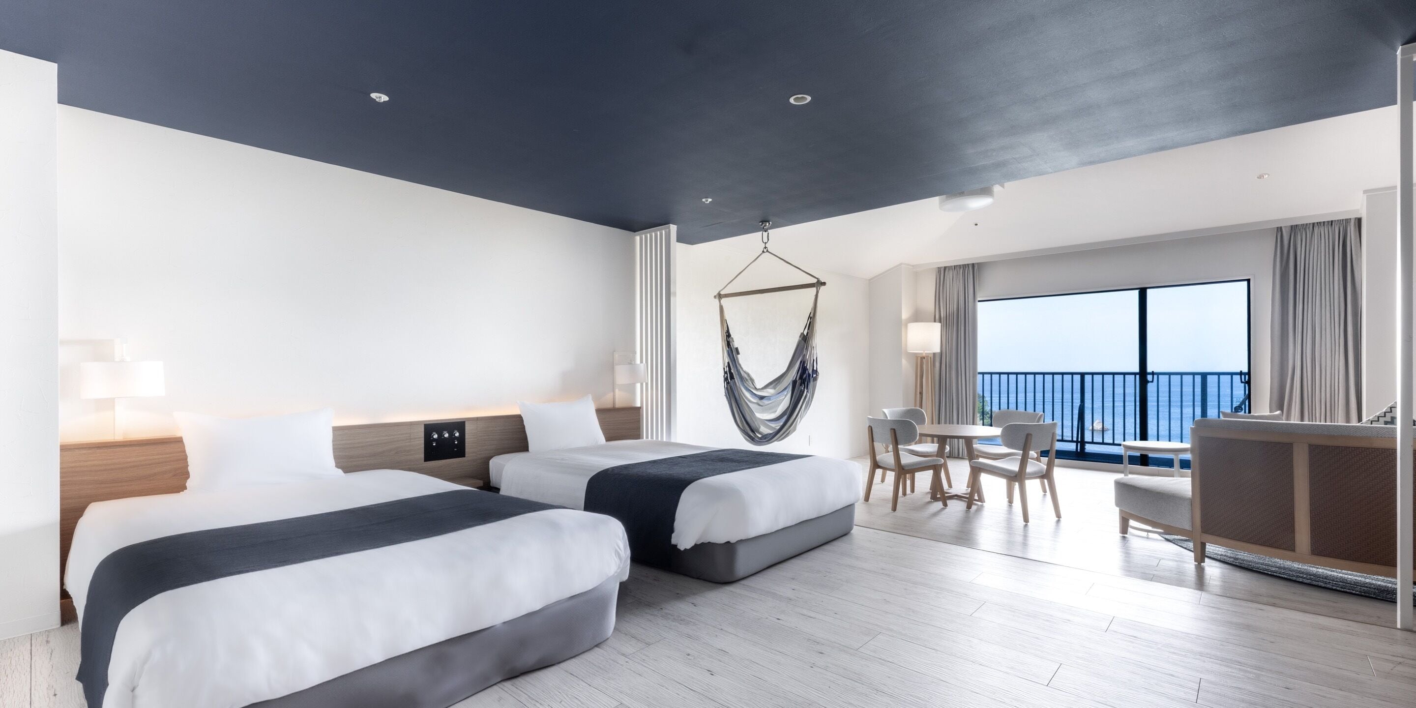 Une chambre d’hôtel avec deux lits, un fauteuil suspendu, un coin repas et un balcon avec vue.