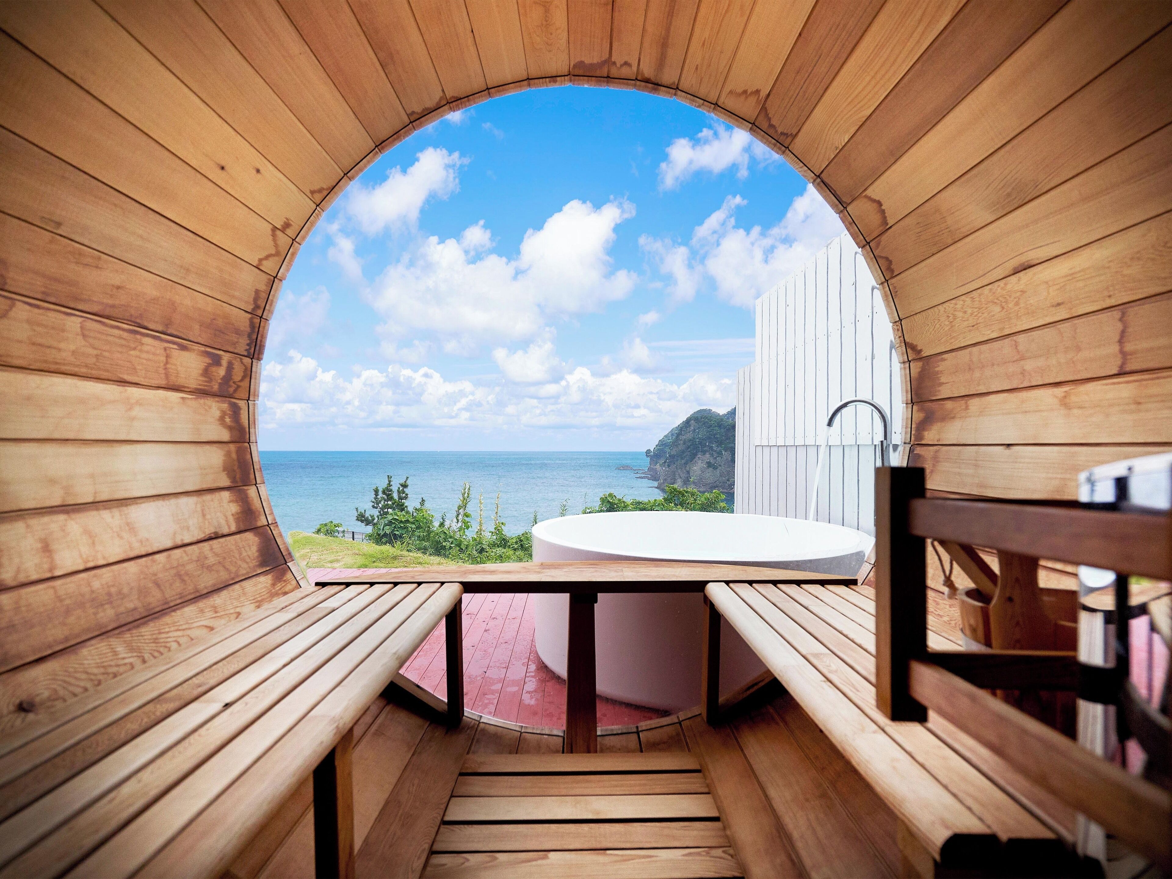 A wooden structure with a circular opening, a white bathtub, and a view of the sea and cliffs.