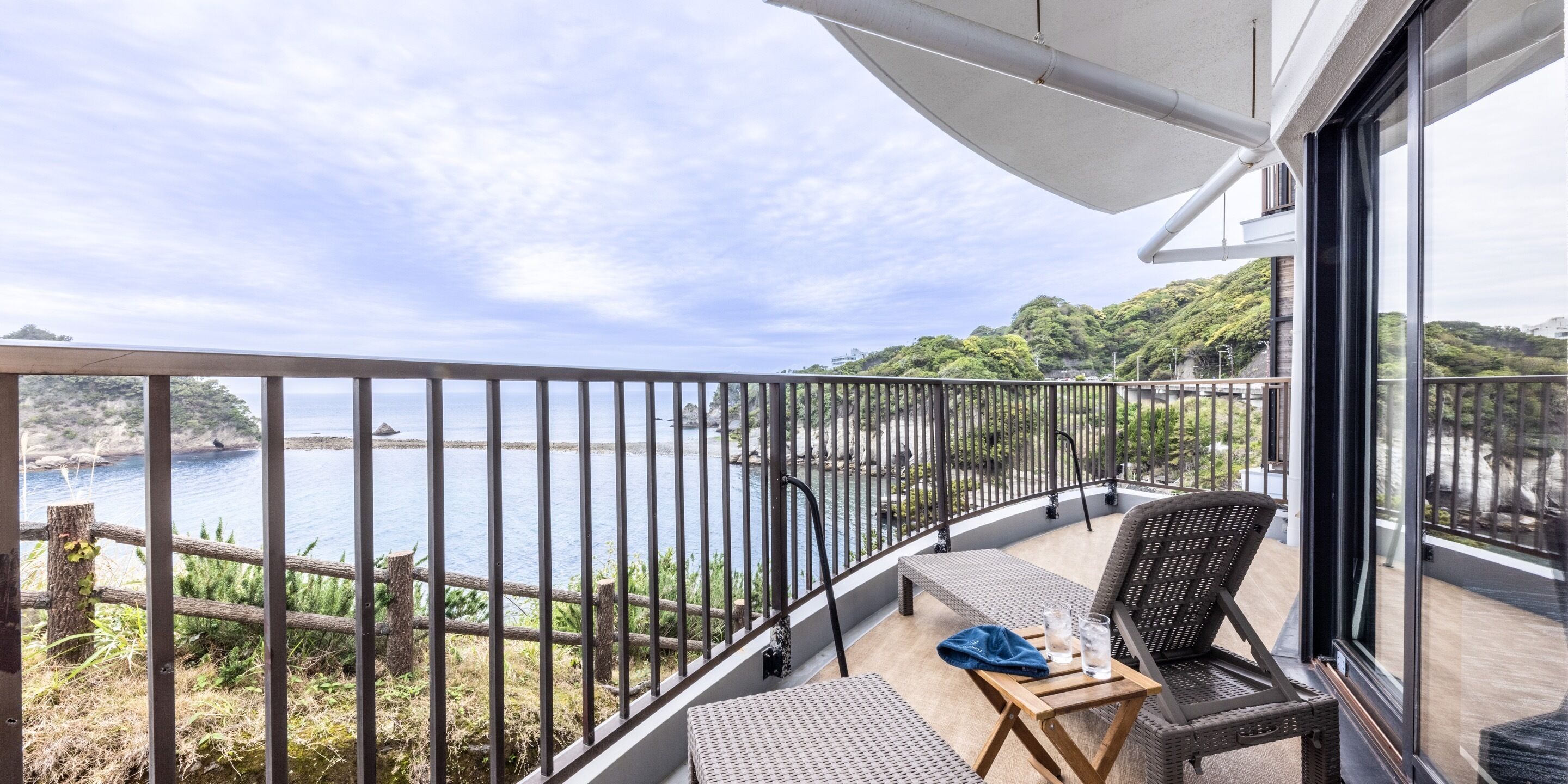 Un balcón con vista al mar, muebles de mimbre y una pequeña mesa con vasos.