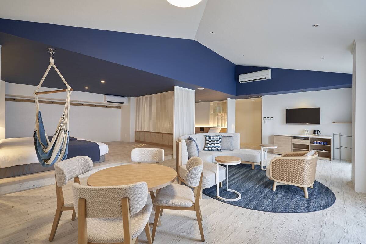 Ein modernes Hotelzimmer mit einem großen Bett, einem Hängekorbstuhl, einer Essecke mit rundem Tisch und Stühlen, einer Couch, einem kleinen Beistelltisch und einem an der Wand montierten Fernseher.