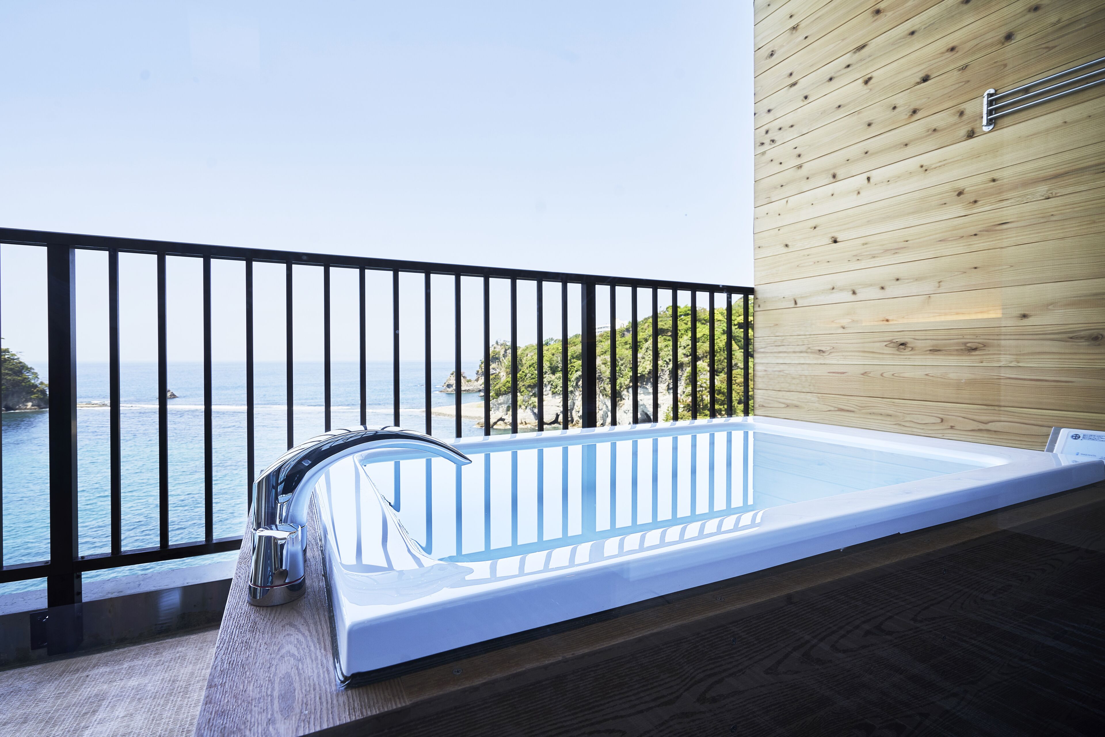 A modern outdoor hot tub with a wooden deck and a black railing, overlooking a body of water and greenery.