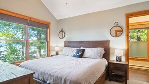 A bedroom with a large bed, two bedside tables, a ceiling fan, and large windows offering a view of trees. - Burt Lake Waterfront Lakehouse (Indian River)