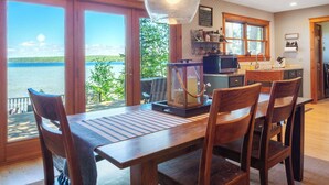 A dining area with a view of a lake, wooden furniture, and a kitchen with a microwave. - Burt Lake Waterfront Lakehouse (Indian River)