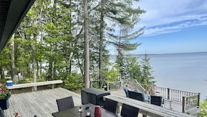 A wooden deck with a table and chairs, overlooking a body of water and surrounded by trees. - Burt Lake Waterfront Lakehouse (Indian River)
