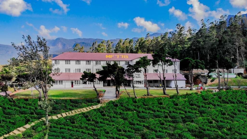 A large building with a red roof and the number 'MF 778' on it, surrounded by lush greenery and mountains in the background.