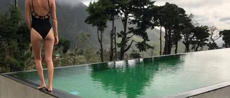 Una persona está de pie al borde de una piscina, con vistas a árboles y montañas al fondo.