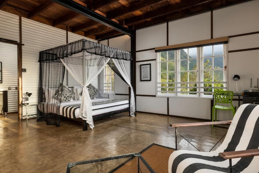 A room with a four-poster bed, a green chair, and a view of greenery through a window.