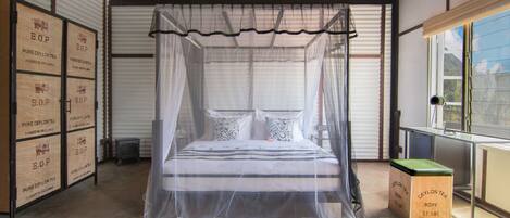 A room with a bed under a canopy, a window with a view, and storage boxes labeled 'B.O.P. PURE CEYLON TEA'.