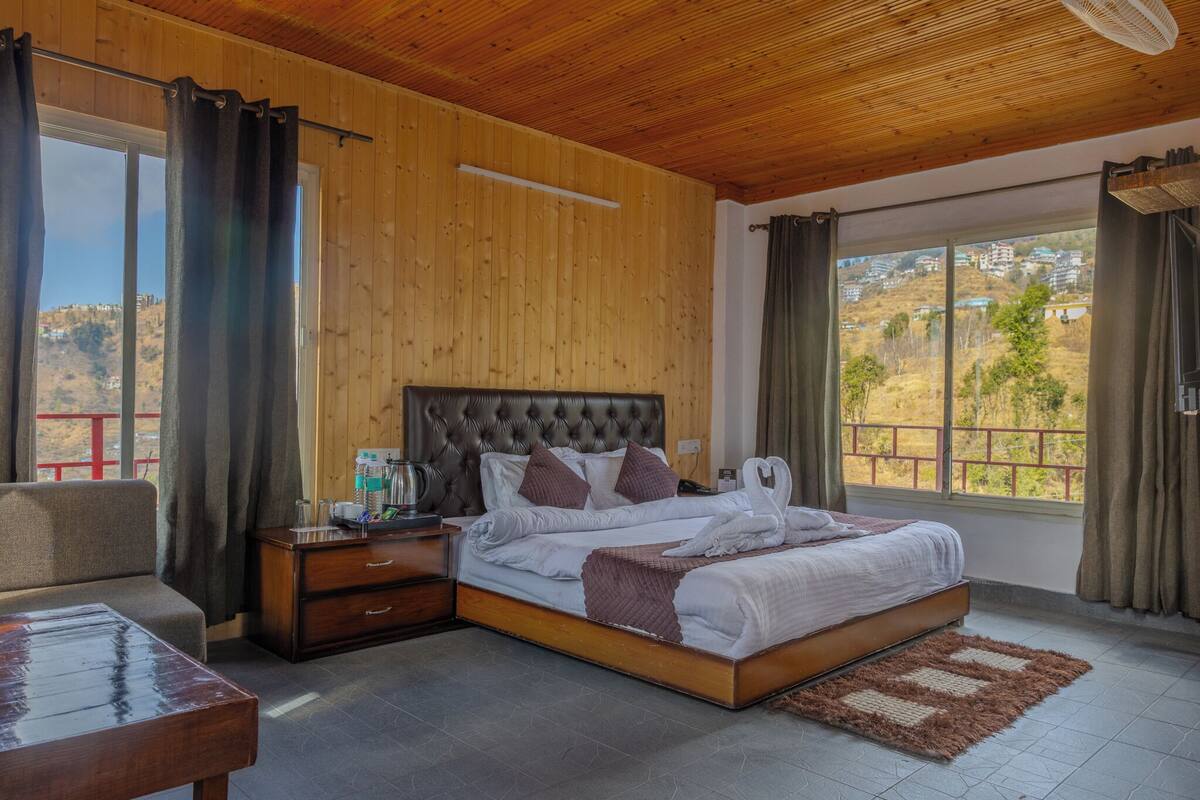 A bedroom with a large bed, wooden walls, a view of the countryside, and a small table with a lamp.