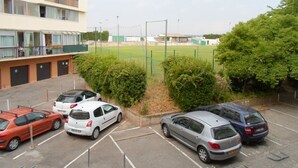Ein Parkplatz mit mehreren Autos, ein Gebäude mit Balkonen und ein Sportplatz im Hintergrund.