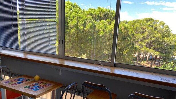 Un aula con un ventanal grande, una mesa de madera con material educativo y tres sillas.