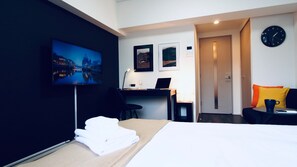 A hotel room with a bed, a desk with a laptop, a TV on the wall, and a clock.