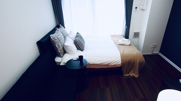 A neatly made bed with a white comforter and a beige throw blanket, a black nightstand with a black cup, and a window with curtains.