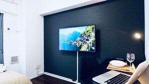 Moderni hotellihuone, jossa on suuri, seinälle asennettu litteänäyttöinen televisio, kannettava tietokone puupöydällä ja viinilasi.
