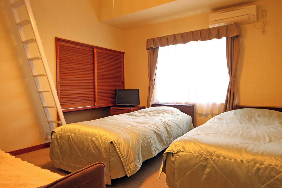 A hotel room with two beds, a TV, a ladder, and a window with curtains.
