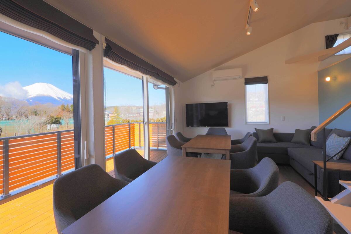 A modern living room with a large wooden table, grey sofas, and a flat-screen TV mounted on the wall.