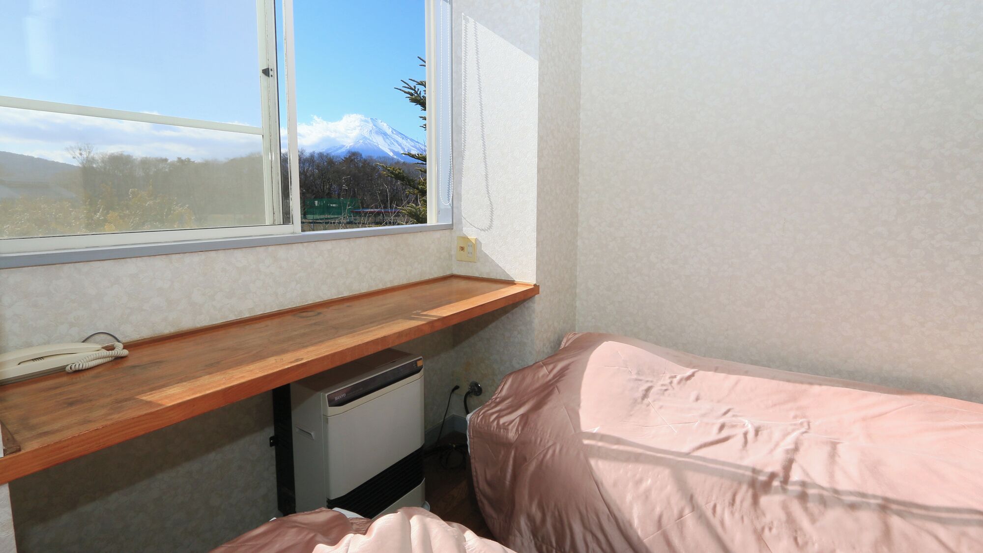 A small room with a wooden desk, a bed, a window with a view of a mountain, and a wall-mounted phone.