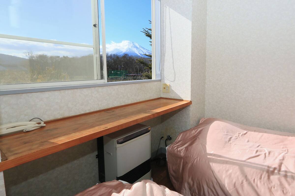 A small room with a wooden desk, a bed, a window with a view of a mountain, and a wall-mounted phone.