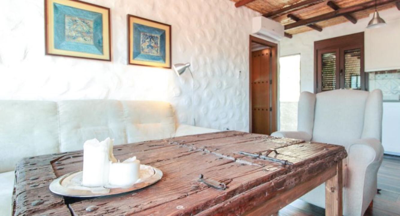 A rustic wooden table with candles, a white cushioned sofa, and a white armchair in a room with textured walls and framed artwork.