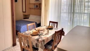 Une pièce de taille modeste comprenant un lit, une table à manger et des chaises. On y trouve également un ventilateur de plafond, une fenêtre avec des rideaux et une étagère remplie de livres.