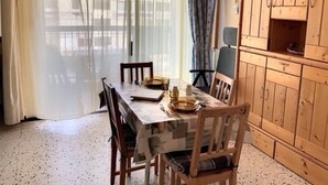 Un coin repas avec une table dressée pour deux personnes, des chaises en bois, un meuble avec des étagères et une fenêtre ornée de rideaux légers.