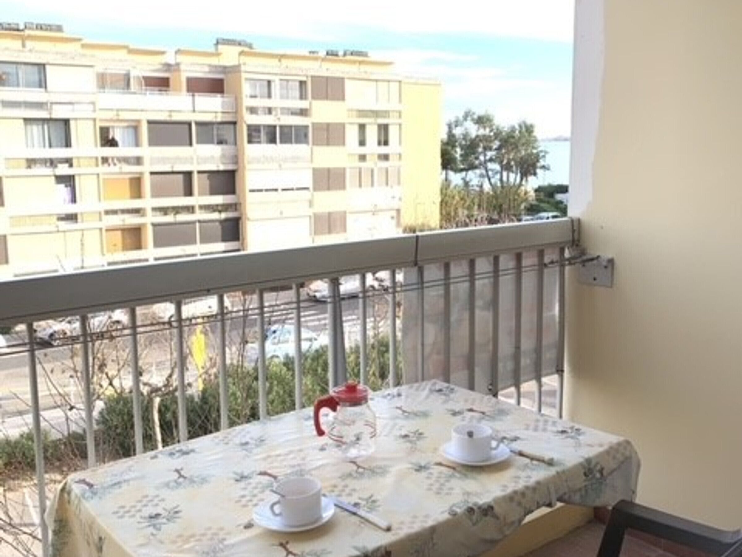 A balcony with a table set for tea, overlooking a building and a parking area.