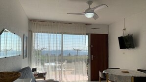Ein Zimmer mit Deckenventilator, einem Esstisch mit Stühlen, einer Couch und einem großen Fenster mit durchsichtigen Vorhängen.
