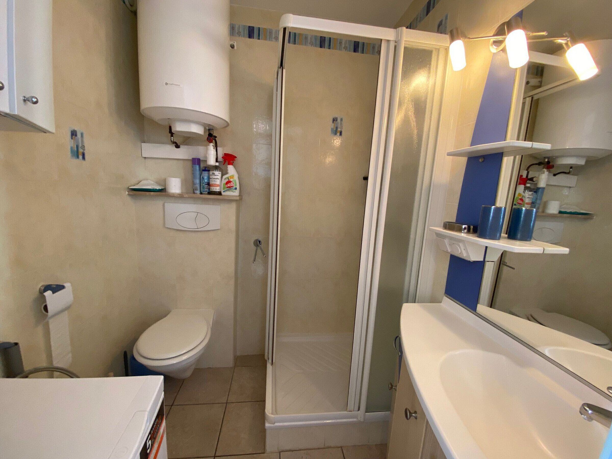 A bathroom with a toilet, shower, and sink. There are shelves with toiletries and a mirror above the sink.