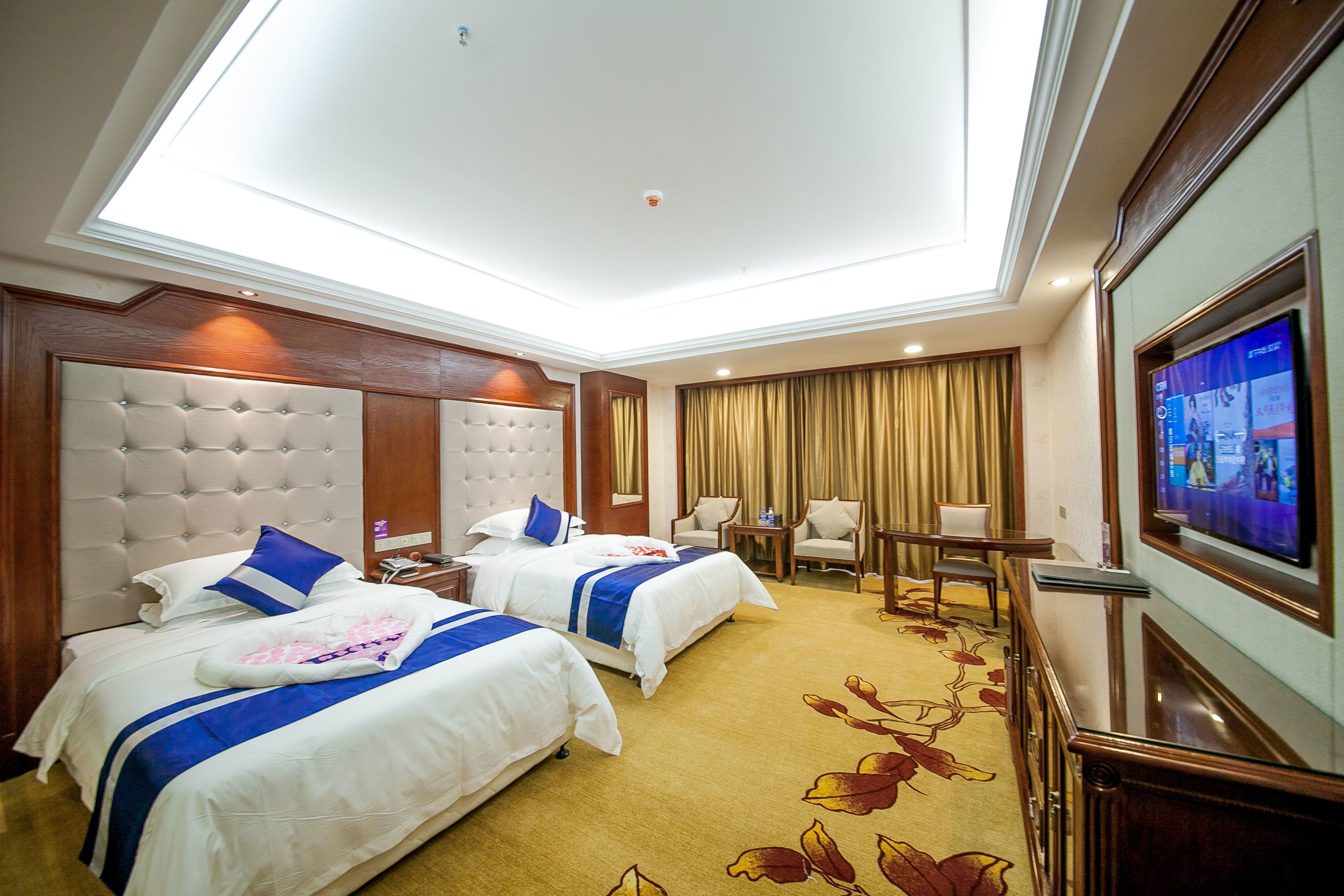 A hotel room with two beds, a television, a desk, and a ceiling light.