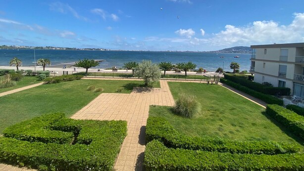 Um jardim com paisagismo e um caminho pavimentado, vista para o mar e um edifício com varandas.