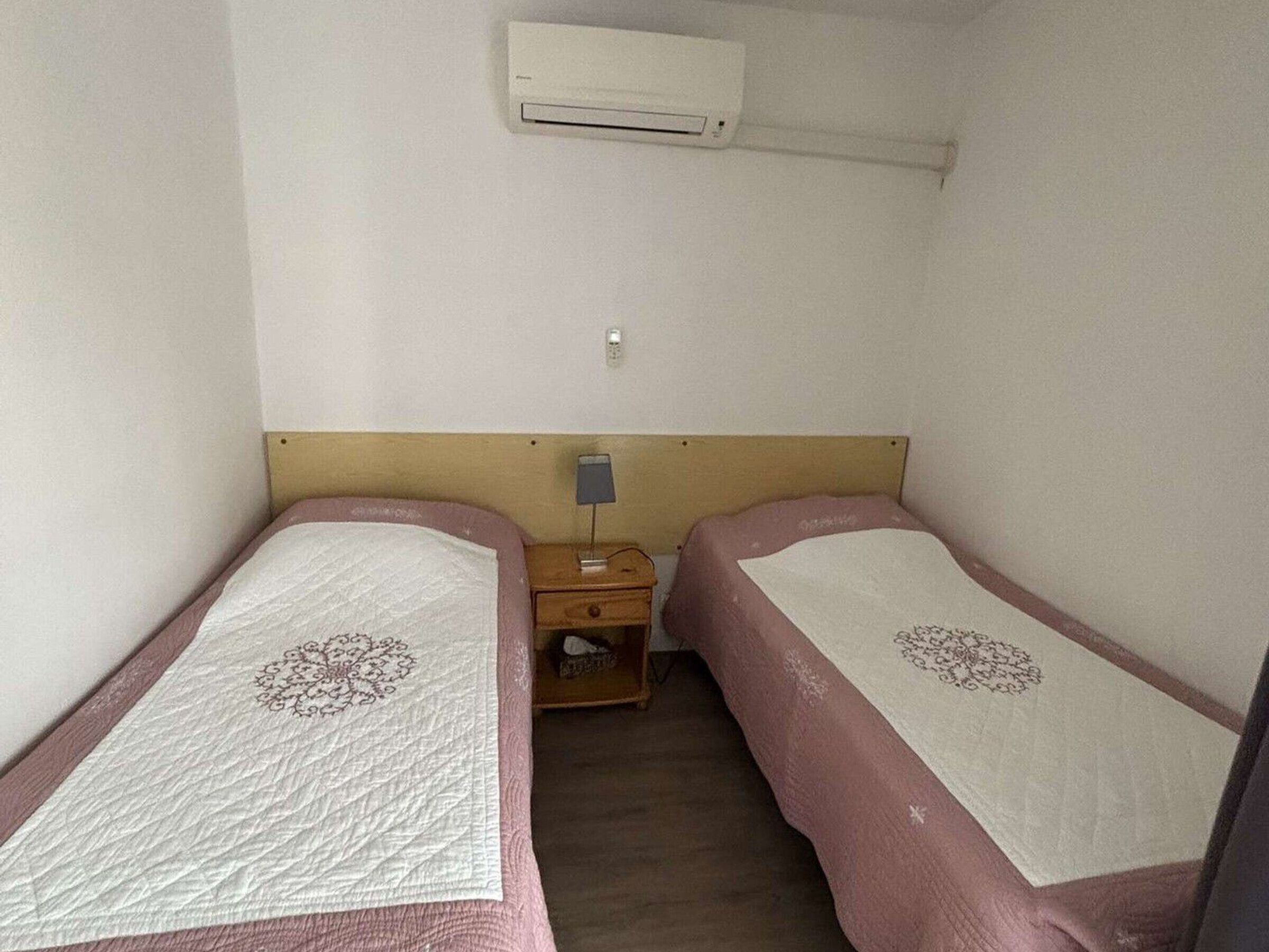 Un alojamiento pequeño y de una sola habitación con dos camas individuales, una mesita de noche, una lámpara y un equipo de aire acondicionado.