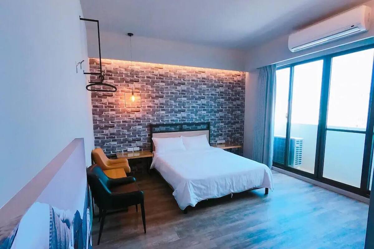 A modern hotel room with a brick wall, a large bed, a desk, and a chair.