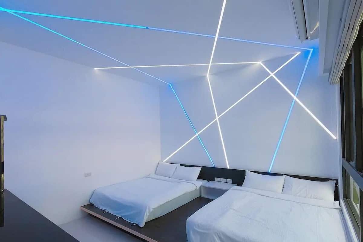 A modern bedroom with two beds, a desk, and geometric light patterns on the ceiling.