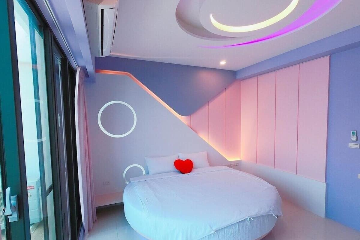 A modern bedroom with a circular ceiling design, a bed with a heart-shaped pillow, and a sliding door on the left.