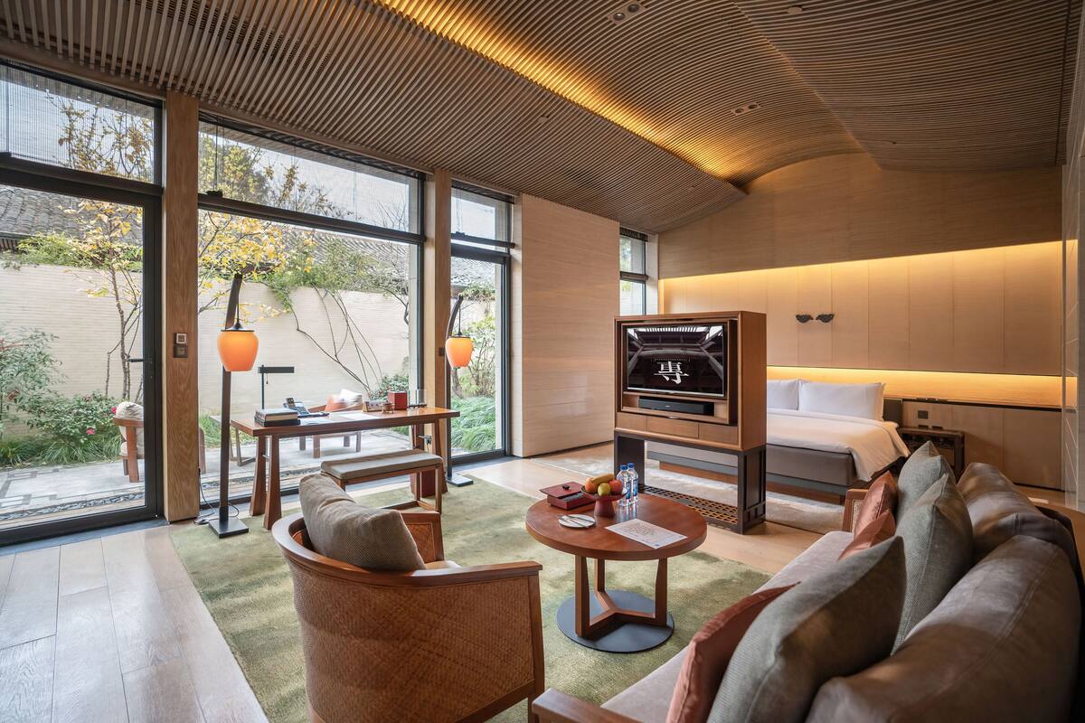 Une chambre d’hôtel moderne avec un grand lit, un canapé, une table basse et un téléviseur. Il y a une fenêtre donnant sur des arbres et un espace patio.