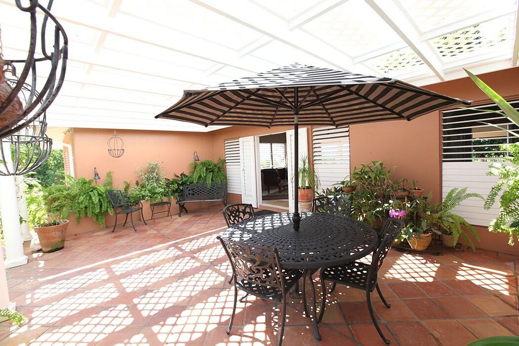 Une terrasse couverte avec une table ronde et des chaises, entourée de plantes en pot et d’un grand parasol.