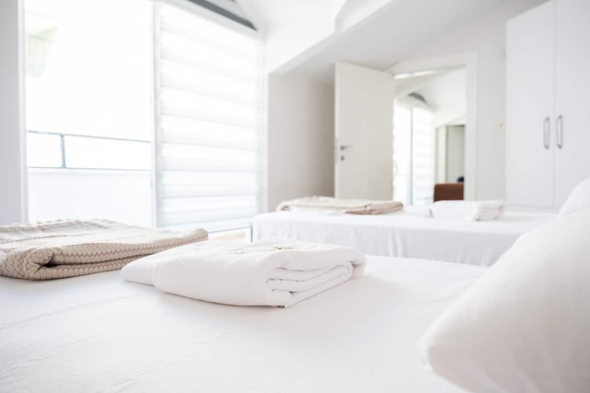 A modern hotel room with a large bed, a neatly made bed with folded towels, a window with blinds, and a white wardrobe.