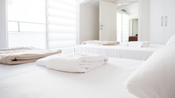 A modern hotel room with a large bed, a neatly made bed with folded towels, a window with blinds, and a white wardrobe.