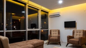 A modern living room with a large window, two armchairs, a sofa, and a flat-screen TV mounted on the wall.