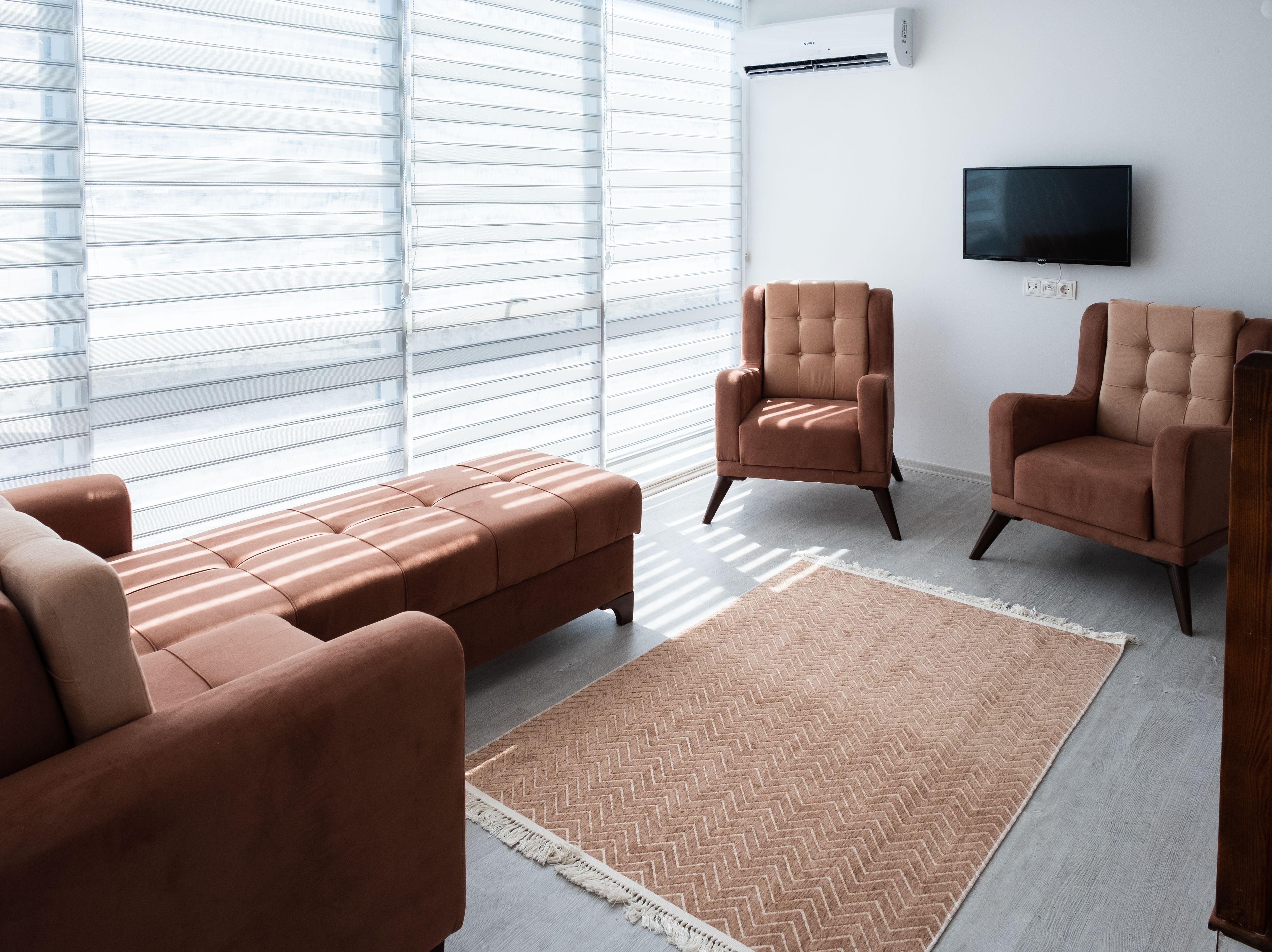 A modern living room with a sectional sofa, two armchairs, a large flat-screen TV, and a large window with blinds.
