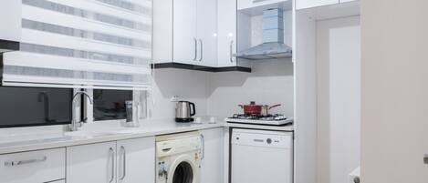 Una cocina moderna con armarios blancos, lavadora integrada, lavavajillas, estufa y ventana con persianas.
