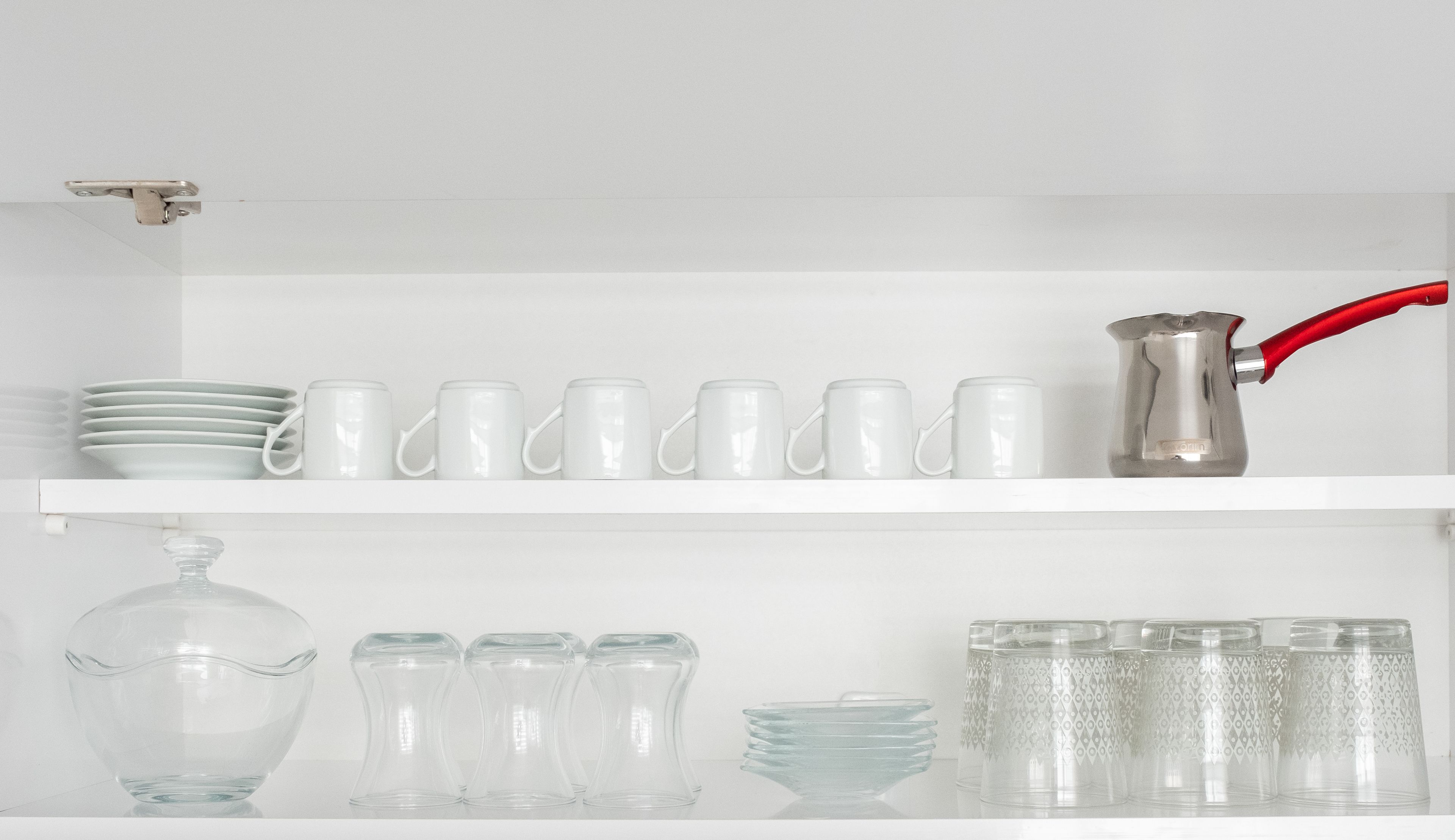 A white shelf with white mugs, plates, a clear glass pitcher, and clear glass cups.