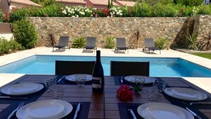 A dining table set for four with plates, cutlery, and a bottle of wine, overlooking a swimming pool with lounge chairs and a stone wall. - Villa, private pool (Oletta)