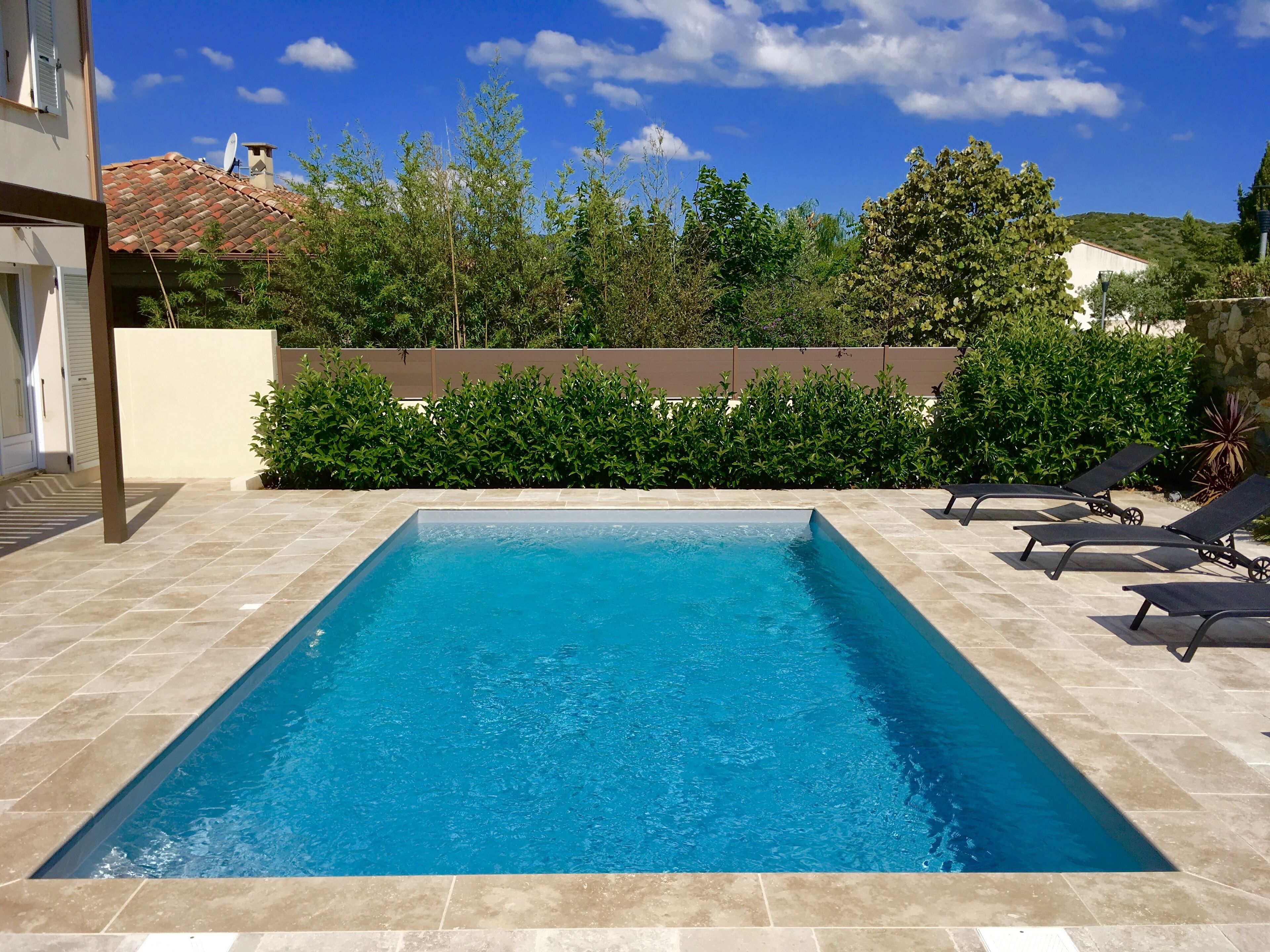A rectangular swimming pool with clear blue water, surrounded by beige tiles. There are lounge chairs along the poolside.